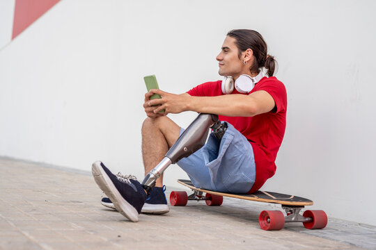 Smiling man with disability sitting on skateboard and holding smart phone