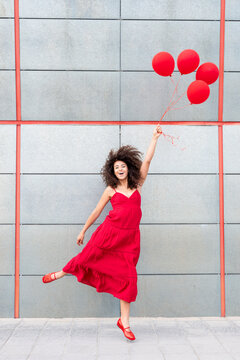 Woman jumping in a red dress with a red balloon by a grey wall