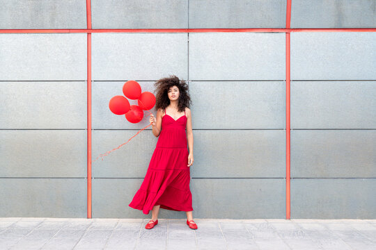 Woman in red dress holding red balloon by grey wall