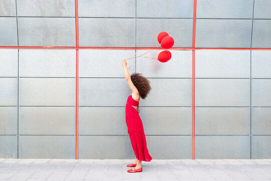 Woman in red dress holding red balloon against grey wall