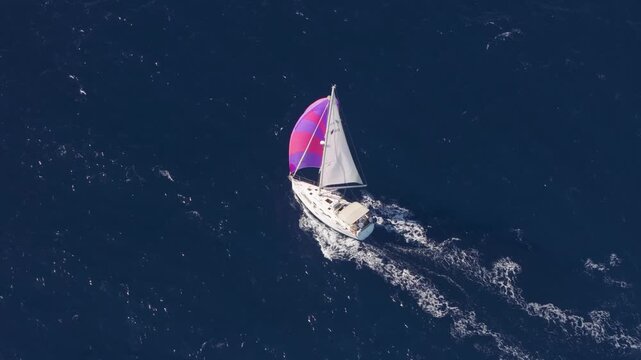 Aerial view of a luxury sailboat cruising on the open sea, featuring a vibrant purple and pink spinnaker sail