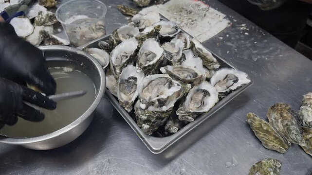 Freshly shucked oysters rest on a metal tray while kitchen staff prepare additional shellfish at a stainless steel workstation. Close-up of professional seafood preparation in a commercial kitchen.
