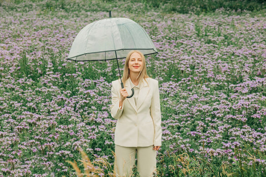 Woman in white suit holds transparent umbrella in phacelia meadow