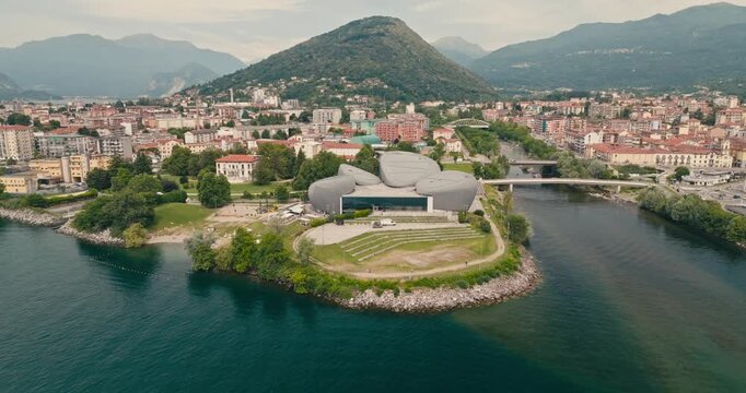 Drone establishing shot approaching Verbania on Lake Maggiore. Turquoise water, orange rooftops, the iconic futuristic theatre and majestic mountains create a stunning Italian lakeside panorama.