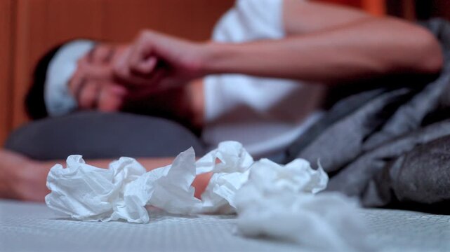 Pile of used tissues near a sleeping ill Asian female with a fever patch. Concept of seasonal virus, health symptoms, and nighttime rest for recovery at home.