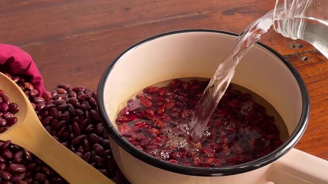 Pouring fresh water from a bottle into an enamel pot with red kidney beans, soaking legumes for cooking close up, organic food. Rustic style