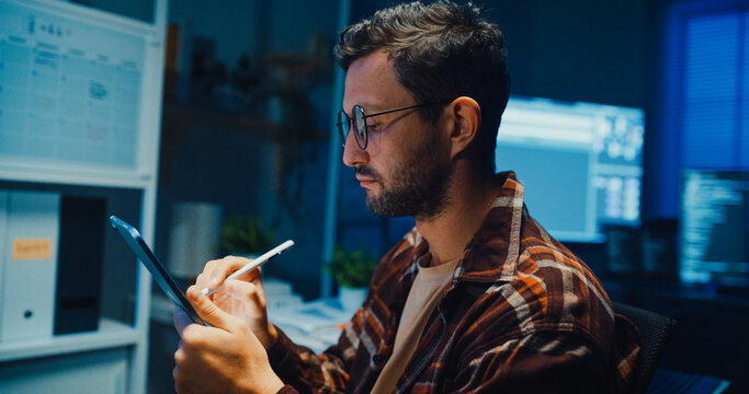 Young Caucasian male programmer use tablet for code design and plan in dark home office at night. AI technology, cybersecurity, software developer, freelance innovation, digital data network concept.