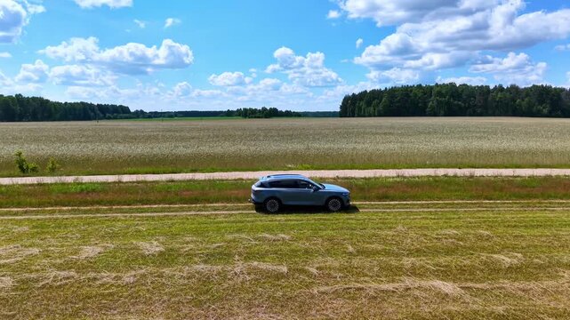 Blue SUV parked on grassy roadside next to expansive green field under blue sky, aerial view