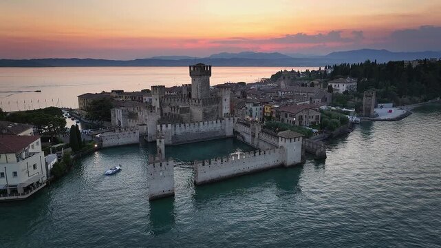 Sirmione, Italy - 4K Flying around of Scaligero Castle (Castello Scaligero di Sirmione) at the comune of Sirmione, located at the lower part of Lake Garda. Sunset, dramatic golden sky and clouds