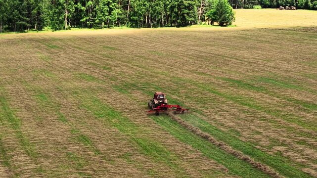 Tractor performing hay cutting on a large grassy field with green trees in the background, aerial view
