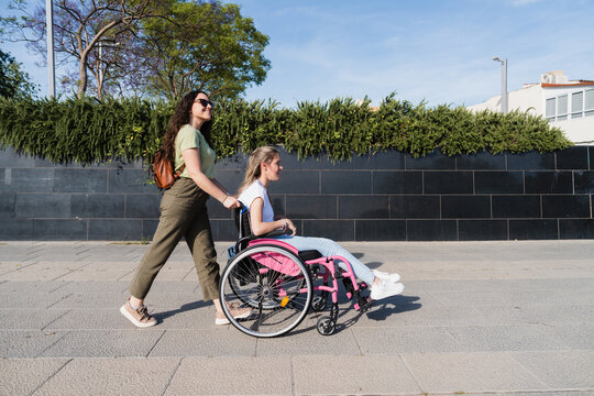 Woman pushes her friend in a pink wheelchair along a sunny sidewalk