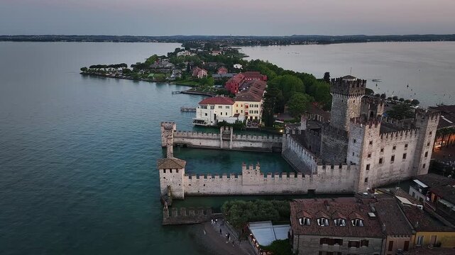 Sirmione, Italy - 4K Flying by Scaligero Castle (Castello Scaligero di Sirmione) at the comune of Sirmione, located at the lower part of Lake Garda. Sunset, dramatic golden sky 
