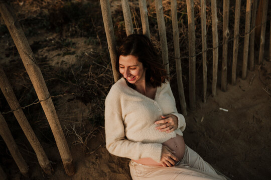Smiling pregnant woman holding her bare baby bump on a sandy beach