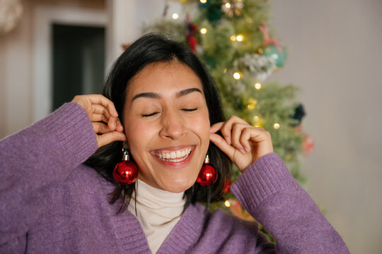 Smiling woman holds red Christmas baubles like earrings at home