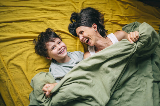 A mother and son laugh together under a green blanket in bed
