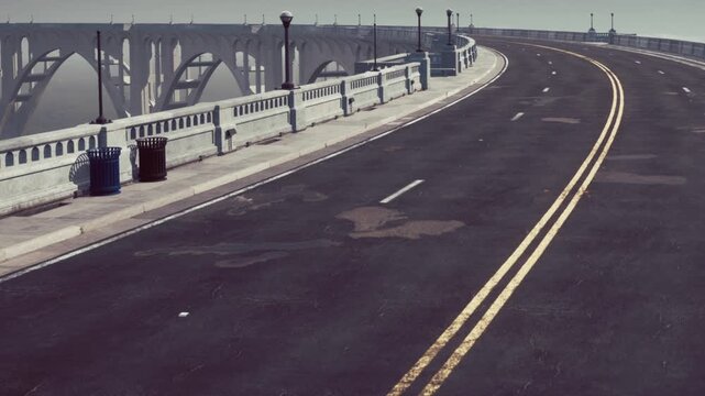 A curved road approaches a large bridge. Street lamps line the edges of the road. The weather appears cloudy with low visibility. The scene shows a spacious view of the structure ahead.