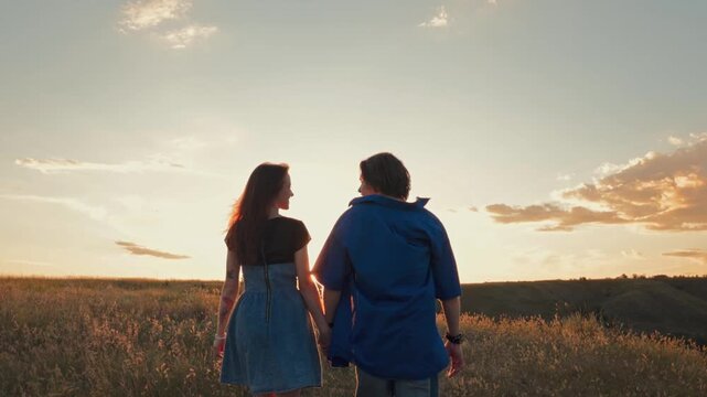 Young couple walking together across meadow at sunset from behind. Concept of romantic getaway, honeymoon travel, relationship, vacation lifestyle and family future.