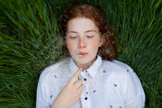 Female with freckles lies in green grass blowing a dandelion