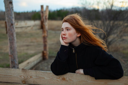 Thoughtful portrait of teenage girl with red hair leaning on a wooden fence outdoors