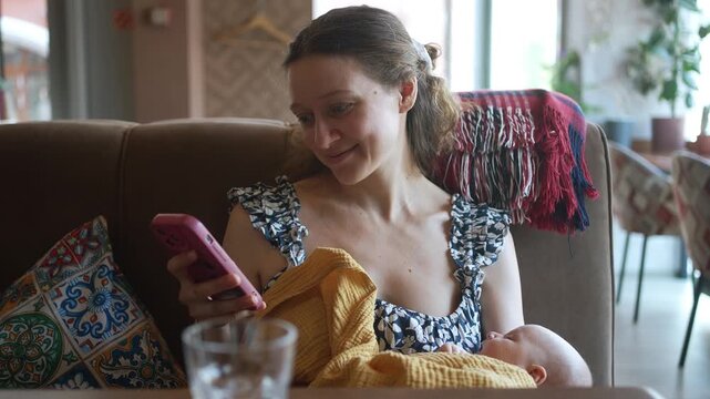 Happy young mother sitting in a cafe, attentively using her smartphone while cradling her sleeping newborn baby in her arms, enjoying a brief personal moment in a public setting