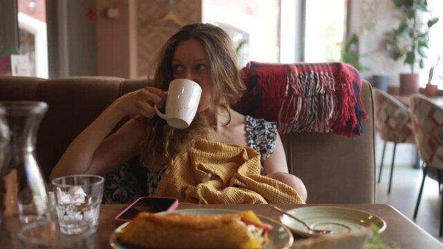 Young mother enjoying a meal in a cafe while breastfeeding her newborn child, she drinks her coffee and tries to finish her food before the baby starts to get restless