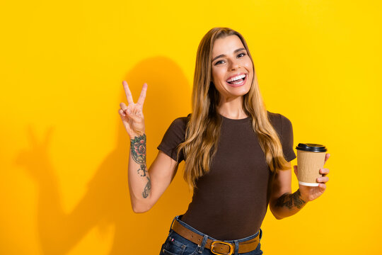 Smiling young woman holding a takeaway coffee cup and flashing a peace sign with visible tattoos, casual style against a bright yellow background