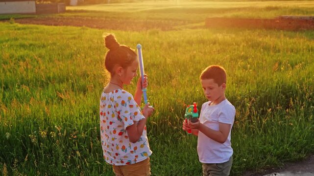 Two children stand in a field enjoying a water spray from a hose. They hold water toys and interact with each other as the sun sets in the background