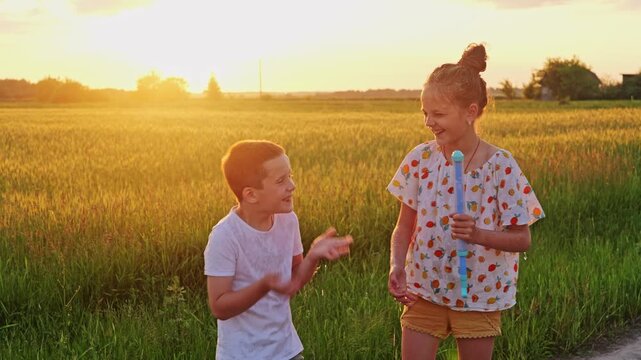 Two children splash each other with water from a hose as the sun sets behind them. They enjoy playing in a green field and laughing together