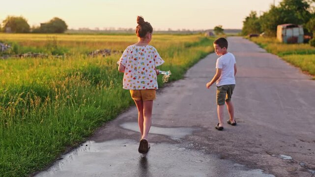 Two children are outside in a field playing with water from a hose as the sun sets in the background. They are enjoying summer and each other's company