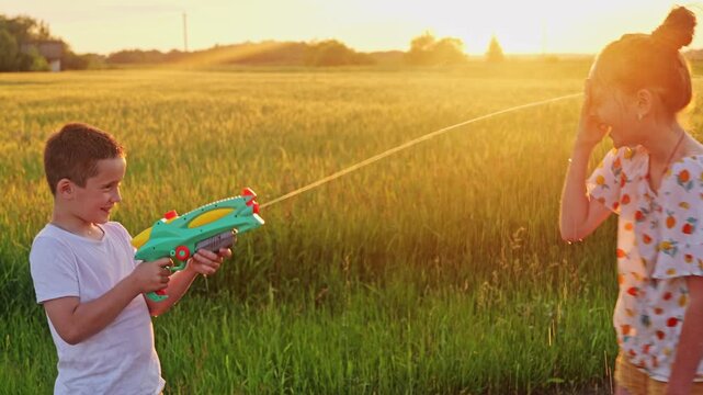 Two children enjoy their time outside as one sprays the other with a water gun in a golden field during sunset in the warm evening light