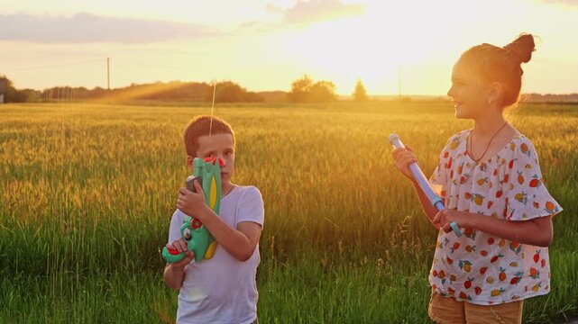 Two children are playing with water guns in a green field. The sun is setting in the background, creating a bright and warm atmosphere. They are smiling and enjoying their time