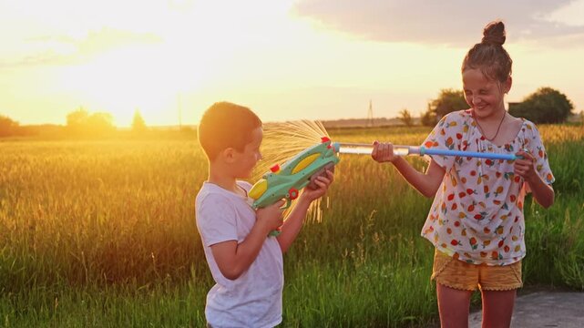 Two children are having fun with water guns in a field. They are spraying each other with water as the sun sets behind them. It is a warm summer evening