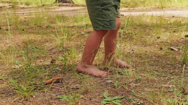 A child walks barefoot on the ground covered with grass and small plants in a forest. The sunlight shines down on the surroundings