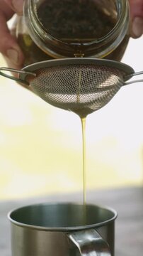 Hands hold glass jar above fine mesh strainer while golden liquid flows into mug beneath steel rim gently
