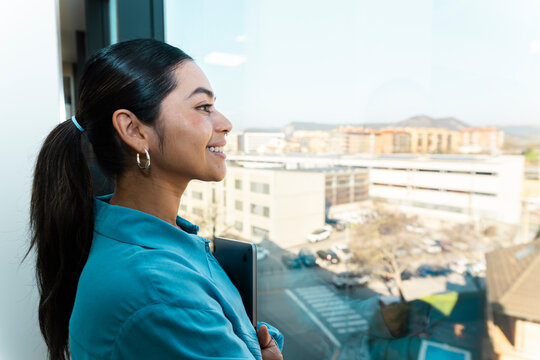 Smiling businesswoman holds laptop and looks through office window
