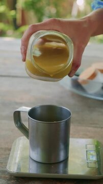 Close view shows hand tilting glass jar, pouring thick golden liquid into metal mug on scale in dim light