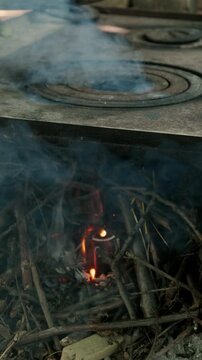 Small flame glows beneath smoke and twigs, revealing worn stove surface, ash, soot, and heat inside room