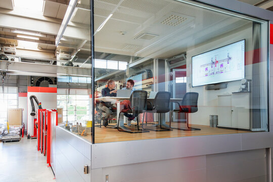 Colleagues discuss a blueprint inside a glass office in a factory