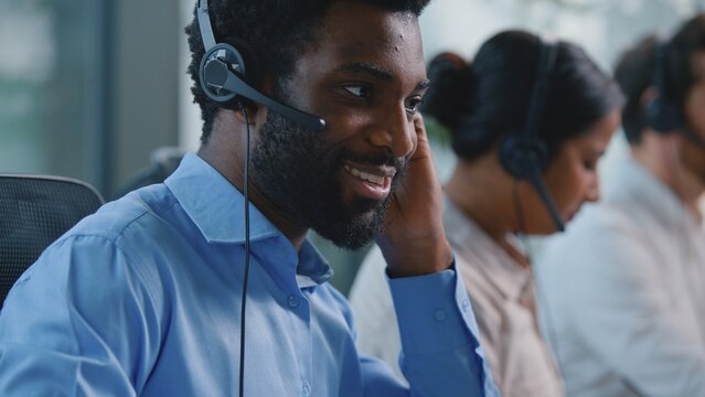 Optimistic smiling African American manager in call center ethnic consultant receptionist talking in headset microphone typing computer keyboard business man carefree talk in callcenter near coworkers