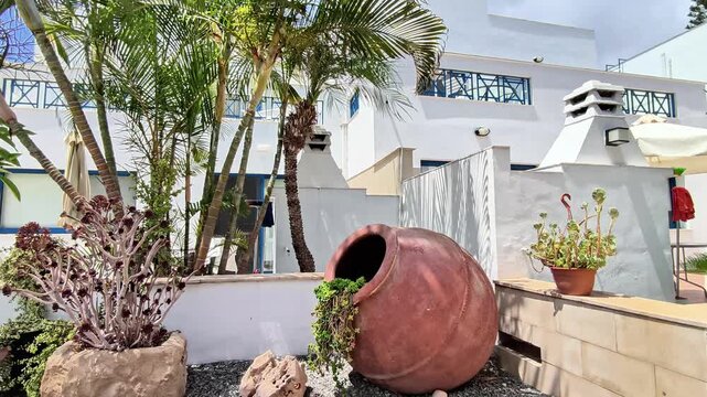 A traditional clay amphora in a landscaped courtyard at a Mediterranean resort in Protaras, Cyprus. Palm fronds sway in the breeze on a sunny summer day, surrounded by succulents and white buildings.