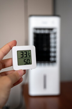Hand holding a digital room thermometer showing high temperature of 33&deg;C against a blurry portable air cooler background. Summer heat and climate control concept