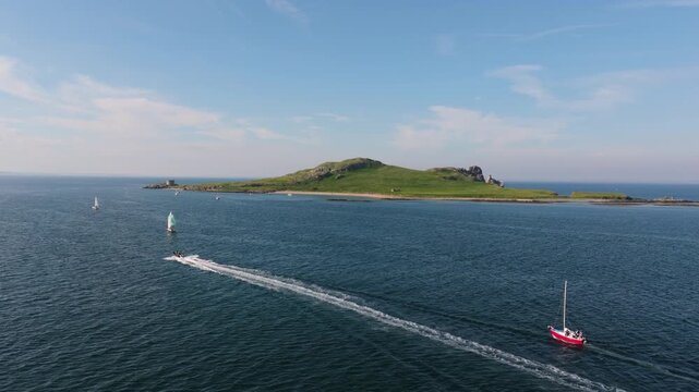 Powerboat and red sailing yacht move across open sea near green Howth headland on a sunny summer day, viewed from above.