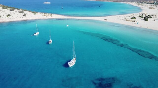 Drone view of sailboats anchored in the crystal-clear turquoise waters of Simos Beach, Elafonisos, Greece, showcasing a pristine sandy lagoon and Mediterranean summer seascape.