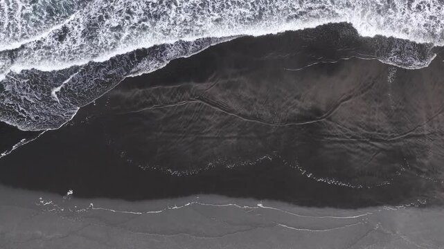 Top-down aerial view of waves washing onto a black sand beach on New Zealand's North Island, creating dramatic ocean textures and natural coastal patterns