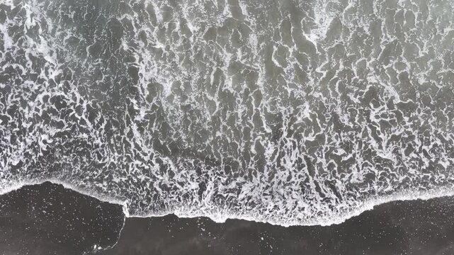 Top-down aerial view of white ocean waves washing across a black sand beach on New Zealand's North Island, creating striking coastal textures and abstract natural patterns.