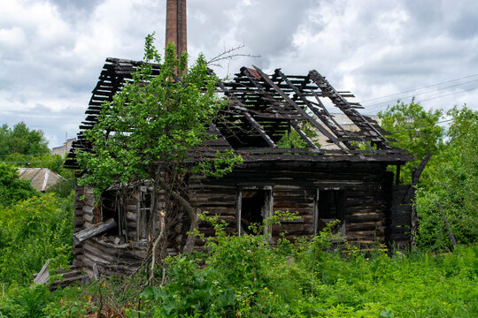 Burnt wooden house ruin with destroyed roof. Abandoned log cabin cottage after fire disaster. Dilapidated rustic structure in overgrown nature scene. Concept of decay and loss.