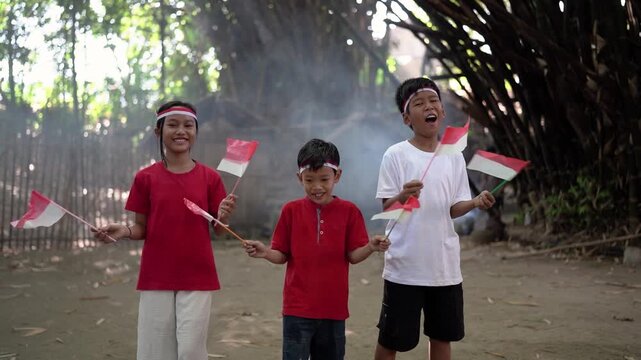 Three children stand on dusty ground in shaded bamboo grove, smiling broadly and raising small red and white flags with joyful energy, soft daylight, and cheerful celebratory mood, with children