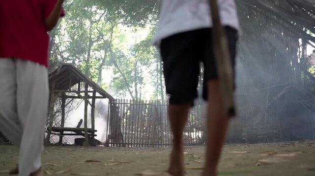 Three children move across dusty ground in rustic forest setting, carrying red and white flags near bamboo hut and wooden gate, with dense trees, soft haze, sunbeams, and lively community mood
