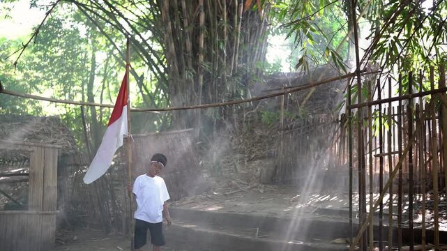 Young boy stands facing away, holding tall bamboo pole beside raised red and white flag near rustic tropical village huts, woven fencing, dense green grove, drifting haze, and soft daylight