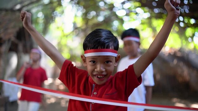 Smiling boy in red shirt stands beside red and white ribbon barrier with both arms raised high, wearing matching headband and showing triumphant energy in sunlit outdoor gathering with blurred